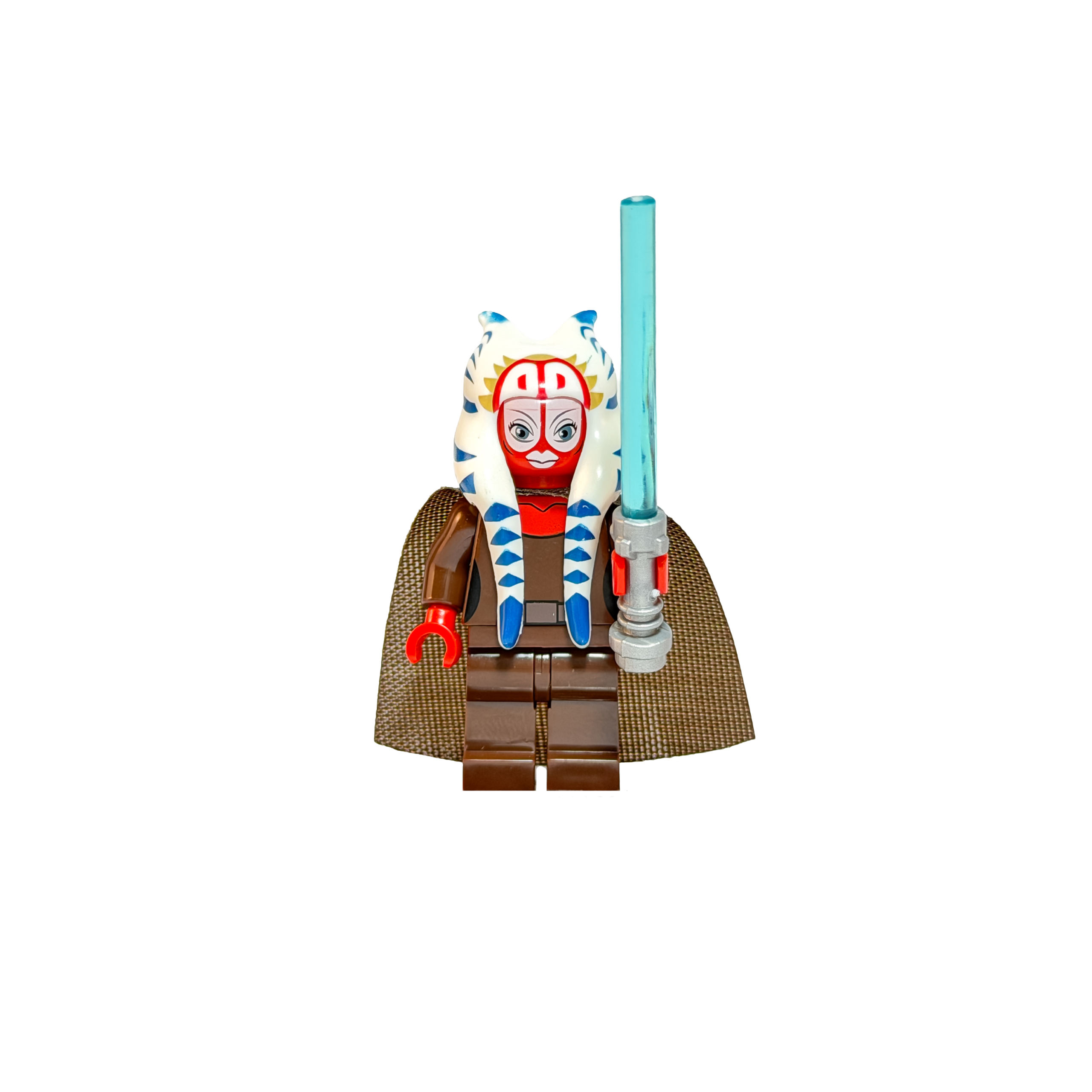 Minifigura de colección de la Maestra Jedi Shaak Ti de Star Wars sobre un fondo negro puro. Es una alienígena de piel roja con un gran tocado blanco con rayas azules que baja por sus hombros. Viste túnicas Jedi marrones y una capa de tela de textura rugosa. Sostiene verticalmente un sable de luz azul brillante con empuñadura plateada.