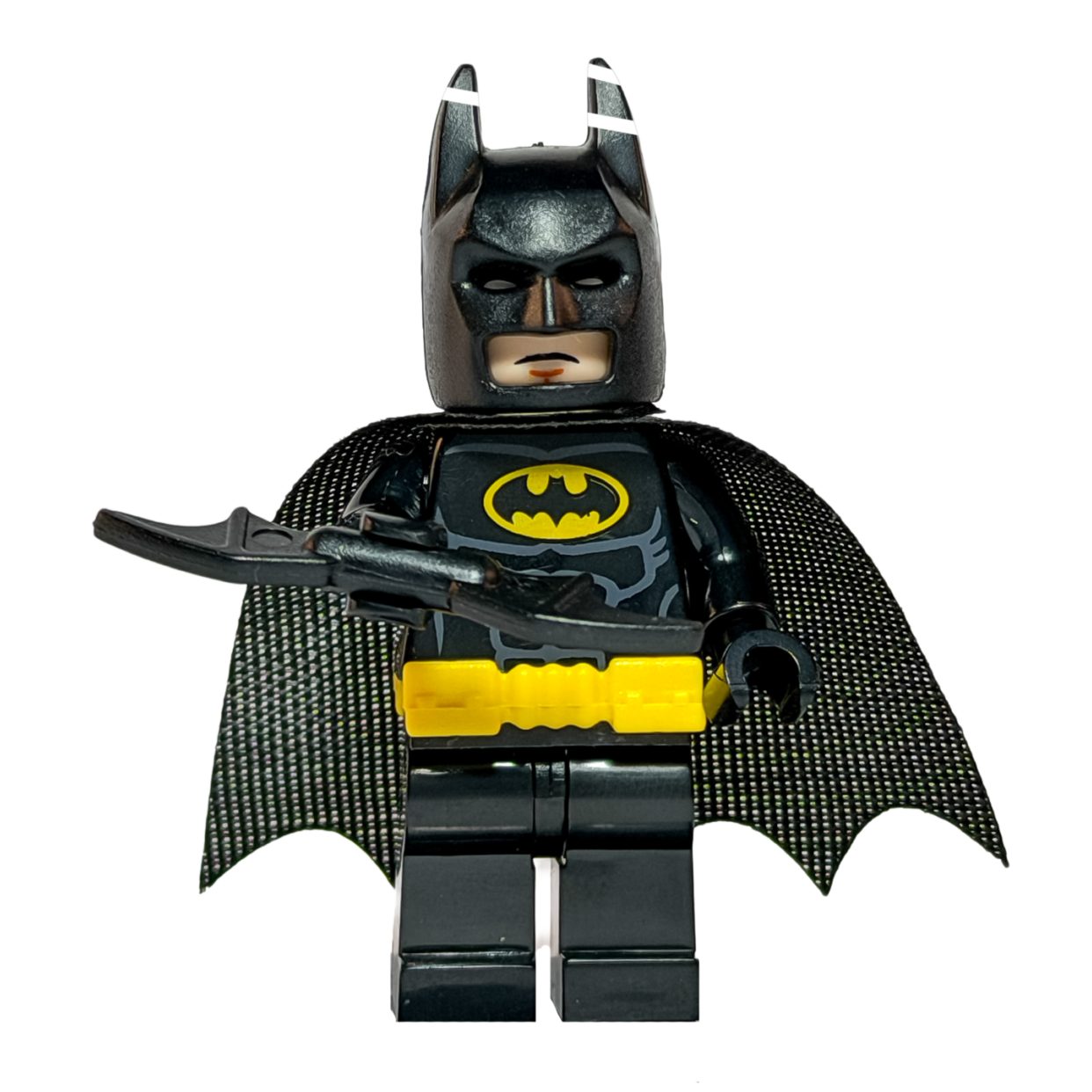 Minifigura de colección de Batman de DC Comics sobre un fondo blanco puro. Viste un traje negro con un cinturón amarillo brillante y el logo del murciélago amarillo en el pecho. Tiene una capa de tela negra y sostiene un Batarang negro en su mano derecha. Está parado sobre una base de exposición negra cuadrada.